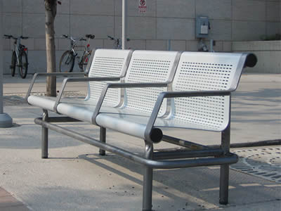 A long chair consisting of three smaller chairs with slotted holes in straight rows is on the street.